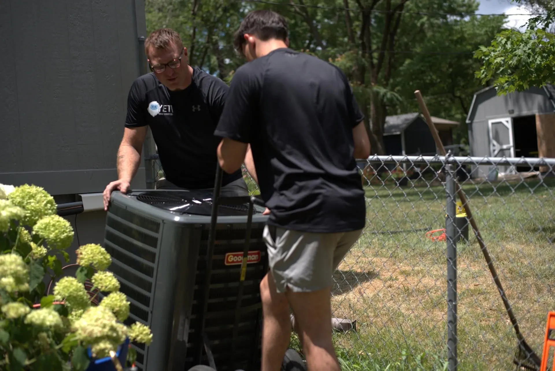 Two technicians setting up an outdoor air conditioning unit beside a backyard fence