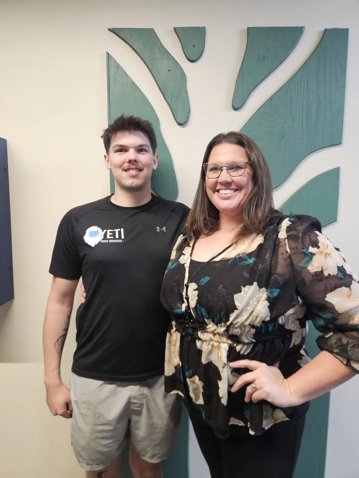 Young technician in a Yeti HVAC shirt standing beside a woman in front of a leaf shaped wall decal